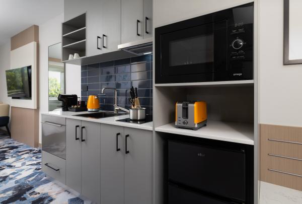 A modern kitchenette at voco Darwin Suites in Darwin, Northern Territory, features grey cabinets, a blue tiled splashback, a microwave, and a mini-fridge. Vibrant orange accents include a stylish kettle and matching toaster. © voco Darwin Suites