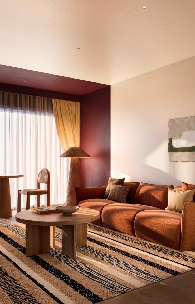 The stylish hotel lounge room at Hyde Perth in Western Australia features a plush terracotta sofa, a striped area rug, a round timber coffee table, and a small dining setting by the window. An open doorway reveals the main bedroom. © Hyde Perth
