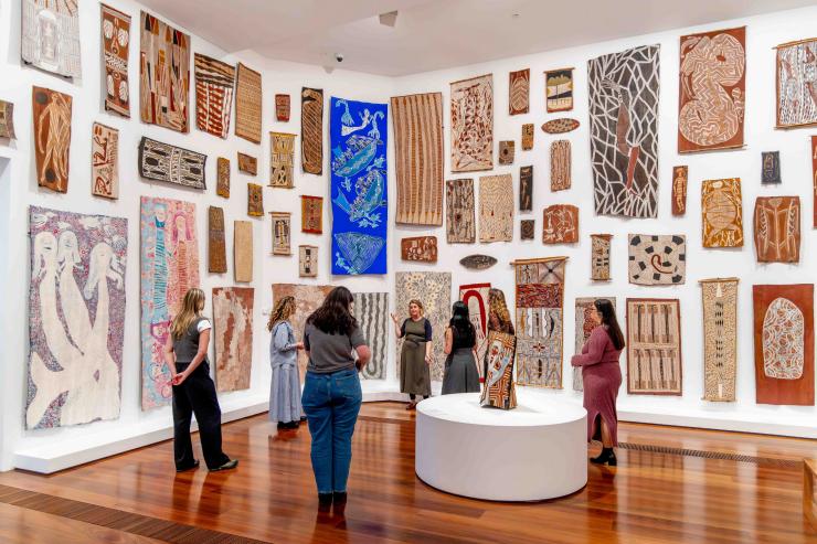 At the National Gallery of Victoria, Melbourne, Victoria, a small group of visitors listens intently to a gallery guide in a bright room. The white walls are densely covered with vibrant Indigenous Australian bark paintings. © National Gallery of Victoria