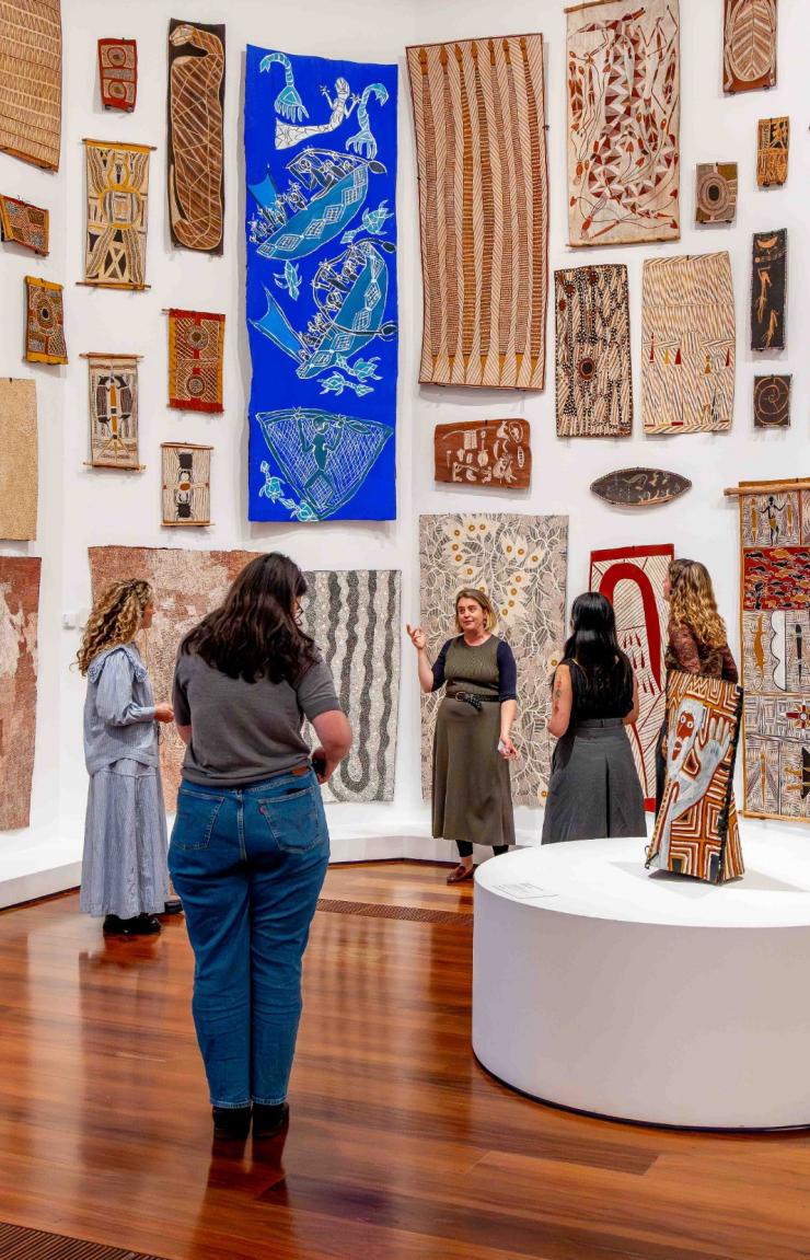At the National Gallery of Victoria, Melbourne, Victoria, a small group of visitors listens intently to a gallery guide in a bright room. The white walls are densely covered with vibrant Indigenous Australian bark paintings. © National Gallery of Victoria