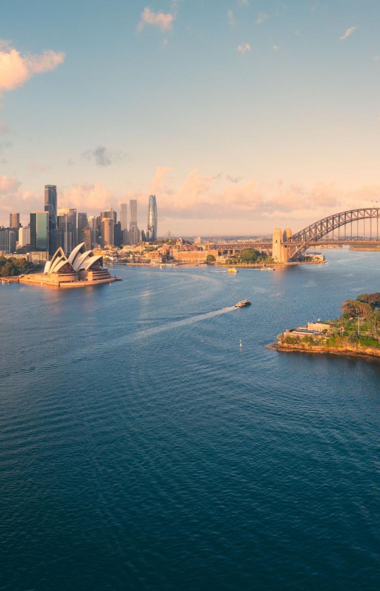 A sweeping panoramic aerial view captures the idyllic and iconic Sydney Harbour in the soft glow of early morning sunshine. The image stretches from the skyscrapers of the CBD and the lush Botanic Gardens across to the famous Sydney Opera House and Harbour Bridge. North Sydney and Kirribilli are clearly visible, basking in the golden light, with the calm harbour waters reflecting the warm hues of the sunrise. © Tourism Australia