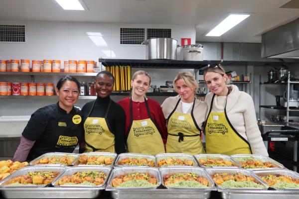 Five women wearing OzHarvest aprons stand at a bench in a commercial kitchen behind trays of prepared food. © OzHarvest