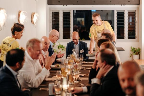 A group of businessmen sit at a dining table at Refettorio OzHarvest Sydney while two OzHarvest team members serve a meal. © OzHarvest
