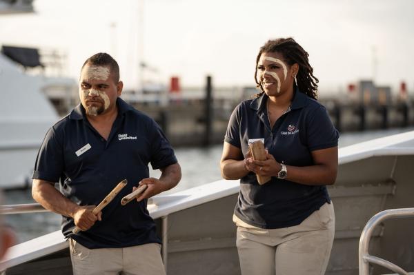 One person holding clapsticks and another with traditional face paint, stand aboard a boat. © Reef Unlimited/GBR Biology