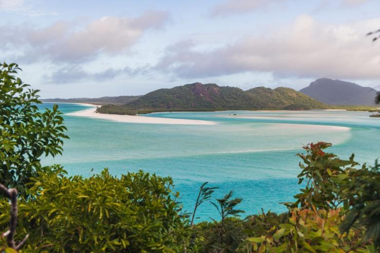 Swirling white sands blend with clear turquoise ocean waters beneath green hills in an elevated view of Hill Inlet, Whitsundays, Queensland © Tourism Australia