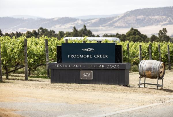 The entrance to Frogmore Creek’s restaurant and cellar door is marked with a barrel and a sign, while sitting behind it are rows of green vines and a rolling landscape, The Precinct Studios © Tourism Australia