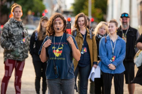 A Blak-led walking tour guide wearing a headset microphone and a t-shirt reading "Sovereignty, Stand United" leads a group of visitors through a city street in Tasmania ©  Jillian Mundy