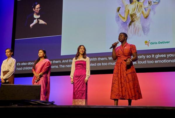 Four young women stand on stage holding microphones at a Girls Deliver session, each wearing attire reflecting her cultural background, while the woman on the right speaks with closed eyes. Behind them, a large screen displays live captioning, a sign language interpreter, and an illustrated graphic alongside the Girls Deliver logo. © Melbourne Convention Bureau