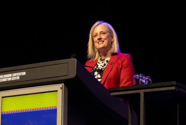 A woman with blonde hair, wearing a red blazer over a black and white floral top, smiles while speaking from a lectern at the Women Deliver 2026 Conference, with the Melbourne Convention and Exhibition Centre and "Hosted by the Oceanic Pacific" branding visible on the lectern panel.