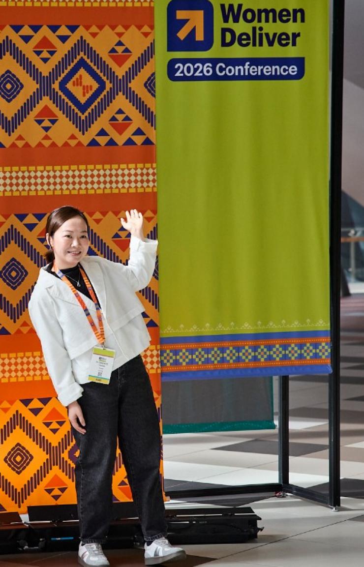 Two attendees pose for a photo at the Women Deliver 2026 Conference, in front of a step-and-repeat display featuring bold yellow, orange and red patterned banners with the phrase "Change calls us here," alongside the Women Deliver 2026 Conference logo. Both women wear lanyards with conference badges, one gesturing toward the banner and the other waving with her hand raised. © Melbourne Convention Bureau