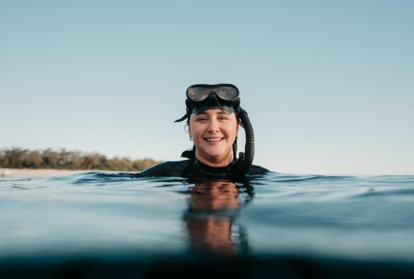 Amy Gash smiling in the ocean while wearing a snorkel with a tree-covered island in the distance, Lady Elliot Island, Southern Great Barrier Reef, Queensland © Tourism Australia