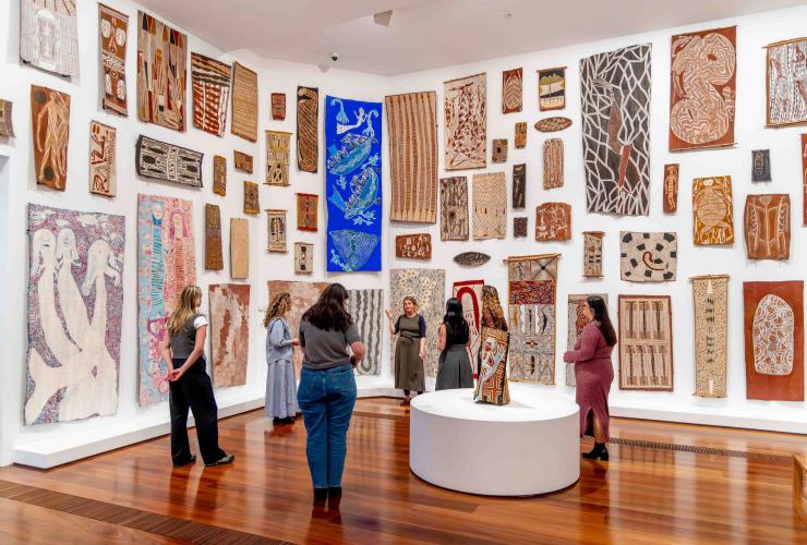 At the National Gallery of Victoria, Melbourne, Victoria, a small group of visitors listens intently to a gallery guide in a bright room. The white walls are densely covered with vibrant Indigenous Australian bark paintings. © National Gallery of Victoria