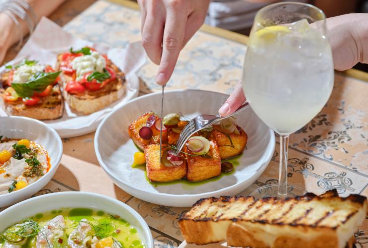 Share plates of bruschetta, hommus, tuna ceviche on a tiled table with a beverage and person cutting into a dish. © Shot by Thom