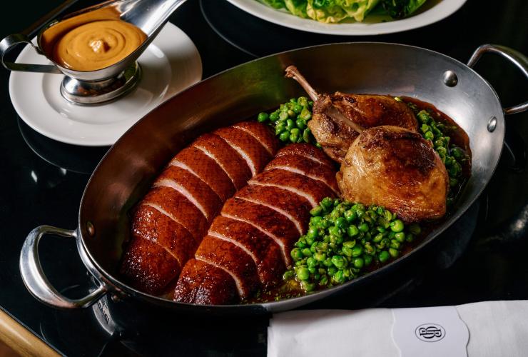 A dish of  grilled duck breast and legs with peas, a sauce dish on a black dining table. © Pete Dillon