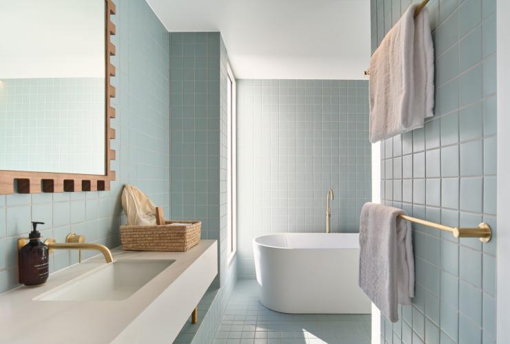 Looking into a bathroom with square blue tiled walls, a sink and mirror on the left hand side  and a free standing bath. © Shot By Thom 
