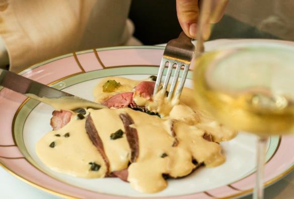 Close up of a person’s hands cutting into a serving of vitello tonnato served on a pink plate next to a glass of wine © Roma Ristorante