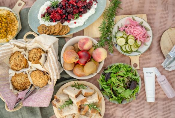 An overhead shot of a picnic blanket and breakfast spread featuring muffins, bread, fruit, pickles and yoghurt. © Beyond Wellness Co & Jurlique