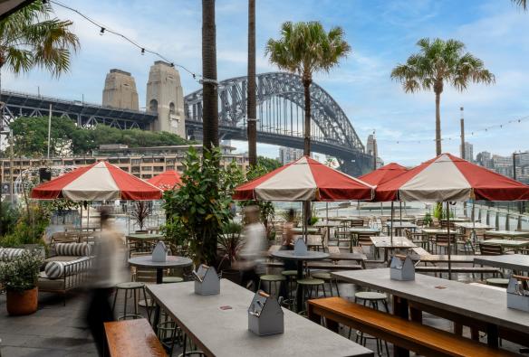 An outdoor pub terrace with large tables and umbrellas sits on the water overlooking the Sydney Harbour Bridge. © The Oriana / Brooke Maxwell