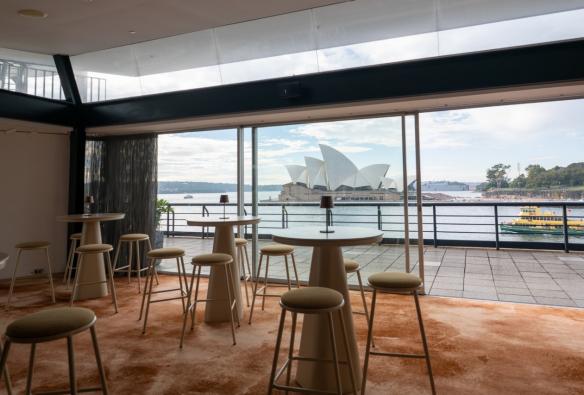 An event room with plush carpet and bar tables is set up near a balcony and large glass sliding doors that look onto the Sydney Opera House. © The Oriana / Brooke Maxwell